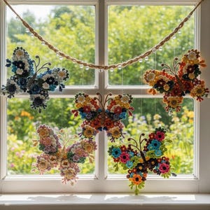 May include: Five colorful, handcrafted butterfly decorations made of flowers hang in front of a window. The butterflies are made of various colors of flowers, including blue, yellow, orange, and pink. A rope with clear beads hangs above the butterflies.