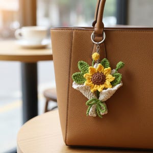 May include: A handmade crochet sunflower bouquet keychain. The keychain features a yellow and brown sunflower with green leaves, wrapped in a white and green crocheted bundle. The keychain is attached to a brown leather handbag.