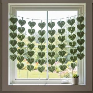 Può includere: Una ghirlanda di cuori verdi all'uncinetto appesa davanti a una finestra. La ghirlanda è composta da più fili di cuori collegati, creando una tenda decorativa. La finestra rivela una vista su un giardino.