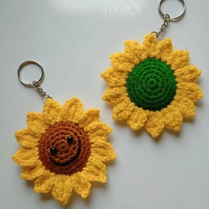 May include: Two handmade crochet sunflower keychains. One has a smiling face with black eyes and a brown center. The other has a green center. Both have yellow petals and silver key rings.