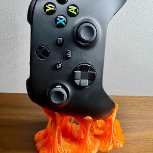 May include: Black Xbox controller resting on an orange, lava-like stand. The controller has colored buttons and directional controls. The stand is a vibrant, textured design, providing a unique display for the gaming accessory.