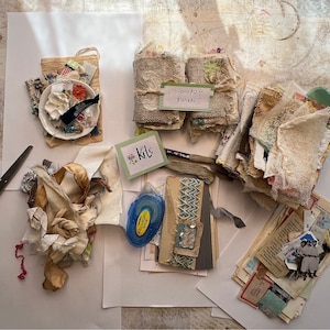 May include: A flat lay of craft supplies for making journals and art projects. The supplies include fabric scraps, paper, ribbon, and a small plate with decorative elements. There are also labels that say "Kits" and "Scrapbook Journals".