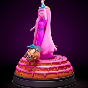 Op de afbeelding: Een kleurrijk beeldje van Princess Bubblegum uit Adventure Time, gekleed in een roze jurk en met een toeter. Het beeldje staat op een drielaagse taart met roze glazuur en hagelslag, met een personage met een schop.