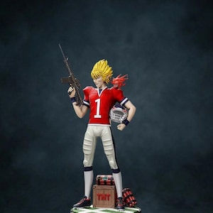 May include: A figurine of a character in a red football jersey with the number 1, holding a rifle and a helmet. The figure has spiky yellow hair and a red dragon-like creature on its shoulder. A wooden crate with "TNT" is at the figure's feet.