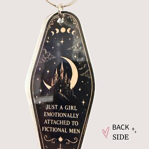 May include: Black and gold keychain with a celestial design featuring a crescent moon, stars, and a castle silhouette. The text reads, "JUST A GIRL EMOTIONALLY ATTACHED TO FICTIONAL MEN."