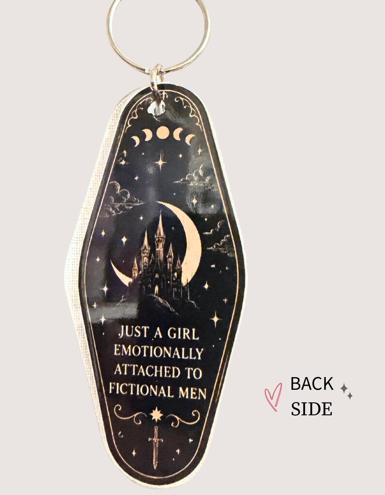 May include: Black and gold keychain with a celestial design featuring a crescent moon, stars, and a castle silhouette. The text reads, "JUST A GIRL EMOTIONALLY ATTACHED TO FICTIONAL MEN."