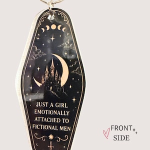 May include: Black and gold keychain with a night sky design, featuring a castle, moon, stars, and the text "JUST A GIRL EMOTIONALLY ATTACHED TO FICTIONAL MEN." The keychain has a silver ring.