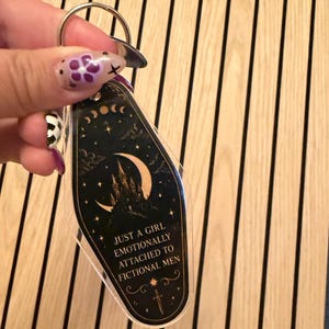 May include: A black and gold keychain with the text "JUST A GIRL EMOTIONALLY ATTACHED TO FICTIONAL MEN." The keychain features a crescent moon, stars, and a castle design. The keychain is held by a silver key ring.