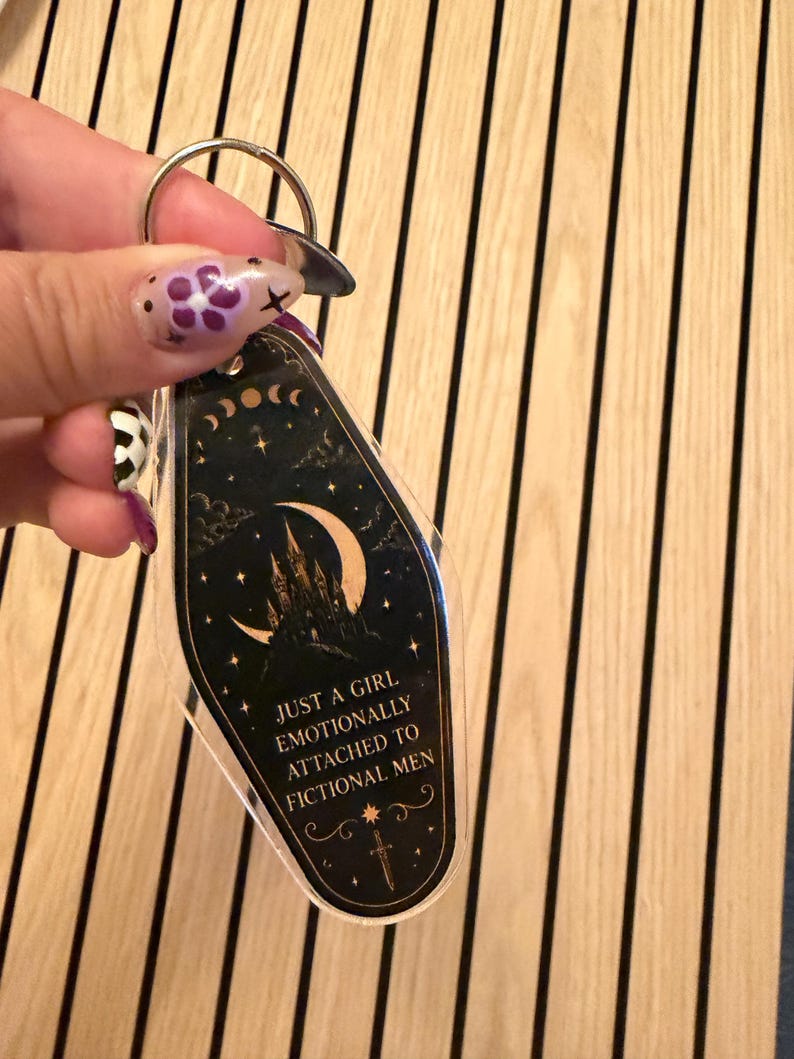 May include: A black and gold keychain with the text "JUST A GIRL EMOTIONALLY ATTACHED TO FICTIONAL MEN." The keychain features a crescent moon, stars, and a castle design. The keychain is held by a silver key ring.