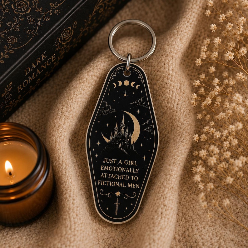 May include: Black and cream-colored keychain with a gothic castle, crescent moon, and star design. The keychain has the text "JUST A GIRL EMOTIONALLY ATTACHED TO FICTIONAL MEN". The keychain is attached to a silver metal ring.