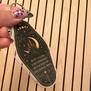 May include: A clear acrylic keychain with a black and gold design. The keychain features a castle, crescent moon, stars, and the text "JUST A GIRL EMOTIONALLY ATTACHED TO FICTIONAL MEN". The keychain is attached to a silver key ring.