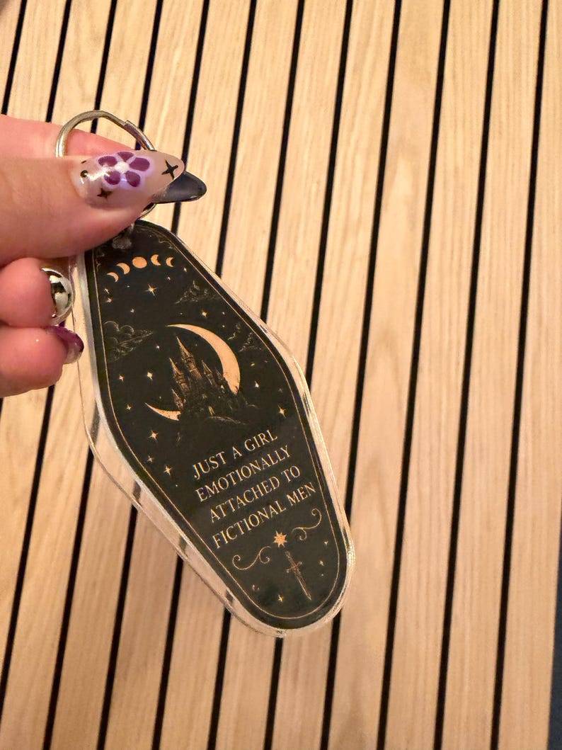 May include: A clear acrylic keychain with a black and gold design. The keychain features a castle, crescent moon, stars, and the text "JUST A GIRL EMOTIONALLY ATTACHED TO FICTIONAL MEN". The keychain is attached to a silver key ring.