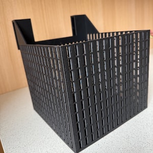 Könnte beinhalten: Ein schwarzer, rechteckiger Aufbewahrungskorb mit einem gitterartigen Muster. Der Korb hat eine quadratische Form mit vertikalen und horizontalen Linien, die kleine rechteckige Öffnungen bilden. Zwei abgewinkelte Stützen ragen vom oberen Rand ab.
