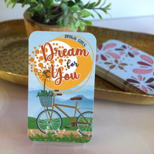 May include: A rectangular card with the text "Dream for You" and a bicycle illustration. The card features a bicycle with a basket of flowers, set against a backdrop of a yellow sun and blue mountains. The words "Brown Girls" are at the top.