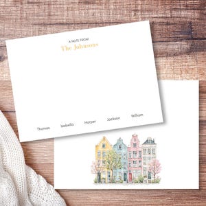 Op de afbeelding: Twee witte rechthoekige briefkaarten. De bovenste kaart bevat de tekst A NOTE FROM The Johnsons in goud en zwart, met de namen Thomas, Isabella, Harper, Jackson en William in het zwart gedrukt. De onderste kaart toont een aquarelillustratie van vier kleurrijke herenhuizen in geel, blauw, roze en grijs met bomen.