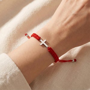 May include: A red woven cord bracelet featuring a central silver-toned metal cross charm. The bracelet has an adjustable sliding knot closure with small silver-toned metal beads at the ends of the cords, creating a simple and functional design.