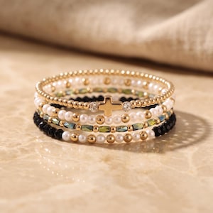May include: A stack of four beaded bracelets featuring a central gold-toned cross charm. The bracelets consist of alternating white pearls, metallic gold spheres, faceted black beads, and iridescent green cylindrical beads, with small clear crystals.