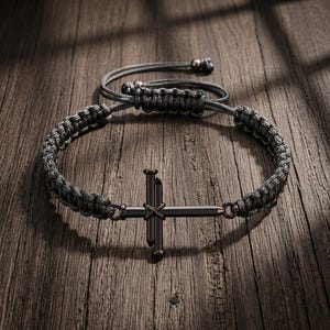 以下が含まれることがあります： ダークグレーの編み込みコードブレスレット。中央には2本の釘のような金属パーツを組み合わせた十字架のチャームが付いています。ブレスレットには調節可能なスライドノット式の留め具があり、コードの端には小さなビーズが2つ付いています。