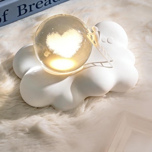 Könnte beinhalten: Ein weißes, wolkenförmiges Nachtlicht mit einer durchsichtigen Glaskugel. Im Inneren der Kugel befindet sich eine leuchtende Herzform. Eine winzige Figur klettert eine Leiter zur Kugel hinauf. Das Nachtlicht steht auf einer flauschigen, weißen Oberfläche.