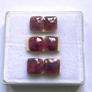 May include: Six faceted, square-cut, reddish-purple gemstones are displayed in a white plastic tray. Each pair of stones is mounted on a small, rectangular piece of beige paper. The gemstones have a polished, reflective surface.