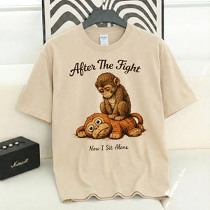 May include: Beige t-shirt with a distressed illustration of two monkeys. One monkey sits on top of the other, with the text "After The Fight Now I Sit Alone" printed above and below the image. The shirt is hanging on a black hanger.