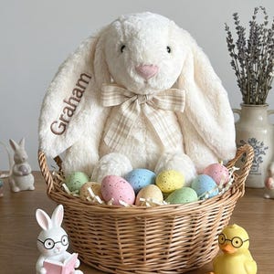 May include: A plush white bunny with the name "Graham" embroidered on its ear sits in a wicker basket filled with colorful speckled Easter eggs. A bow tie adorns the bunny's neck. Decorative figurines of a reading bunny and a duckling with glasses are in front.