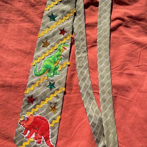 Peut inclure: Une cravate vert olive clair avec un motif à carreaux subtil, ornée d'un écusson T-Rex vert, d'un écusson Tricératops rouge, d'une bordure en croquet jaune et de plusieurs petites décorations en forme d'étoile rouge, verte et dorée.