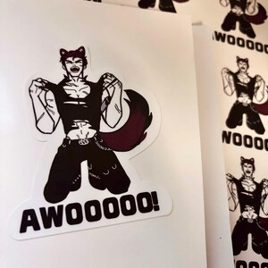 May include: A die-cut sticker featuring a stylised illustration of a character with cat ears, wearing a black sleeveless top and trousers, with a large dark tail. The character is shown in a dynamic pose with the bold black text AWOOOOO! below.