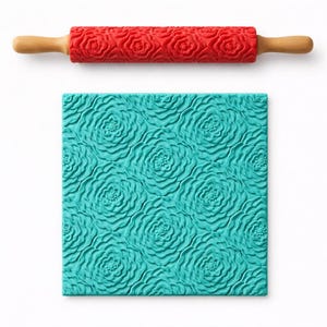 May include: A red rolling pin with a floral pattern and wooden handles is shown above a teal, floral-patterned square. The rolling pin is designed for baking and creating textured surfaces on dough or fondant.