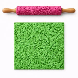 Op de afbeelding: Een roze deegroller met een bloemenpatroon en houten handvatten wordt getoond boven een groen, bladerenpatroon vierkant. De deegroller is ongeveer 30 cm lang. Het groene vierkant heeft een gedetailleerd reliëfontwerp van bladeren en planten.