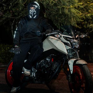 May include: A black hooded sweatshirt featuring a white lightning bolt graphic on the hood and face mask, paired with black pants and black gloves, positioned on a white and black motorcycle with red wheels against a dark outdoor background.