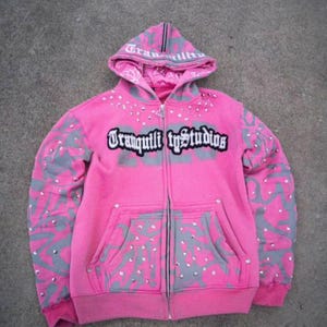 Könnte beinhalten: Ein pinkfarbener Kapuzenpullover mit Reißverschluss und dem Text "Tranquility Studios" auf der Brust. Der Hoodie hat graue Akzente an Ärmeln und Kapuze sowie dekorative Verzierungen. Er hat eine Vordertasche und eine Kapuze mit Kordelzug.