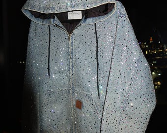 May include: Light blue denim jacket with a hood, embellished with numerous sparkling rhinestones. The jacket features a front zipper, a kangaroo pocket, and a brown leather patch. The hood is lined with a dark fabric, and the cuffs and hem are ribbed.