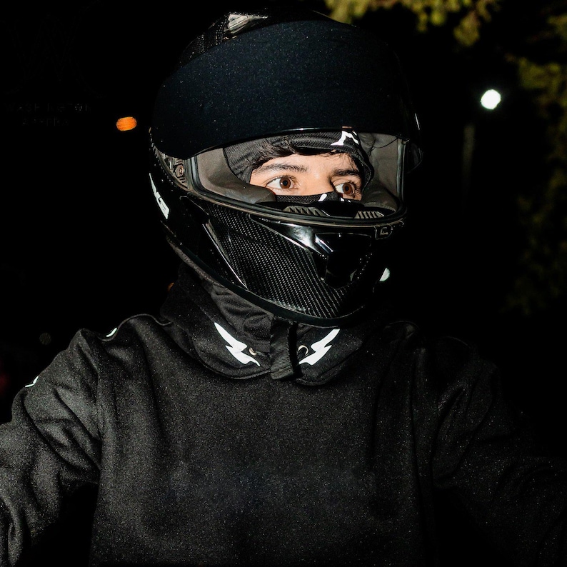 May include: A black full-face motorcycle helmet with a carbon fiber texture on the chin guard and a clear visor. The helmet is worn over a black balaclava and a black long-sleeve shirt featuring white lightning bolt graphics on the collar area.