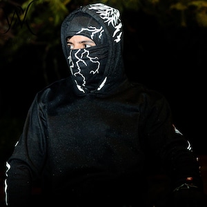 May include: A black hooded sweatshirt featuring a white lightning bolt pattern on the hood and a matching black face mask with a white lightning bolt design. The garment includes long sleeves with white lightning accents along the outer arms.