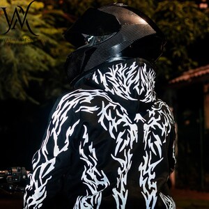 May include: A black hoodie featuring a white, jagged, flame-like graphic pattern across the back and hood. The garment is worn with a black carbon-fiber textured motorcycle helmet that has a dark, tinted visor and a chin guard.