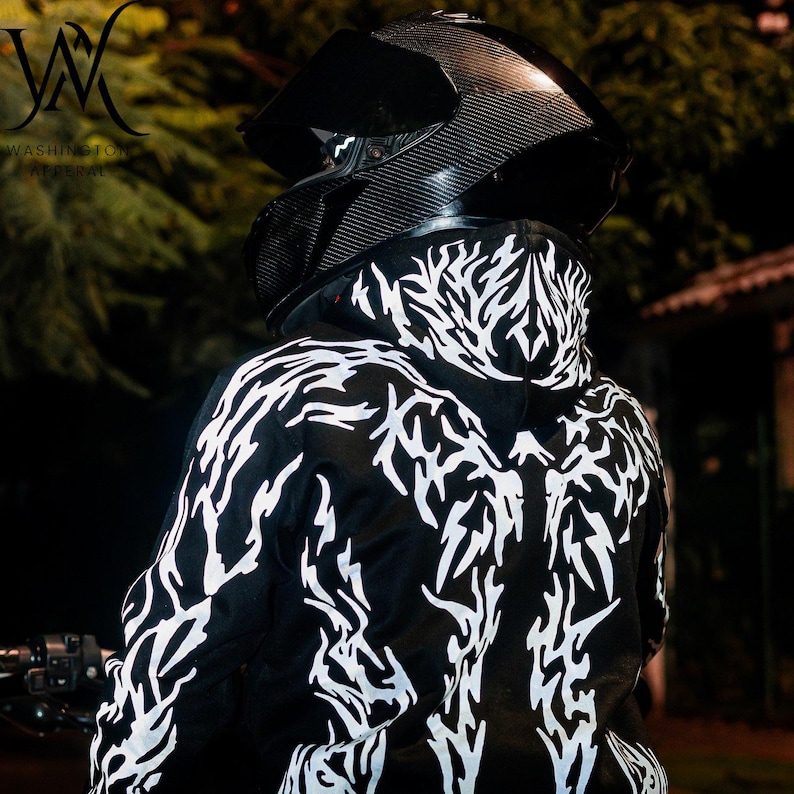 May include: A black hoodie featuring a white, jagged, flame-like graphic pattern across the back and hood. The garment is worn with a black carbon-fiber textured motorcycle helmet that has a dark, tinted visor and a chin guard.