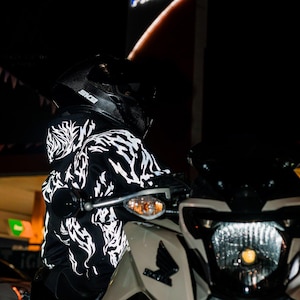 May include: A black hoodie featuring a white, jagged, flame-like graphic pattern across the fabric. The hoodie is worn with a black carbon-fiber style motorcycle helmet with the text SKB visible on the side, positioned on a white motorcycle.
