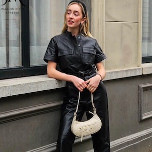 May include: A black short-sleeved jumpsuit with a button-up front, two chest pockets, and a matching belt. The jumpsuit is paired with a cream-colored crescent-shaped shoulder bag featuring a front zipper pocket and gold-tone hardware.