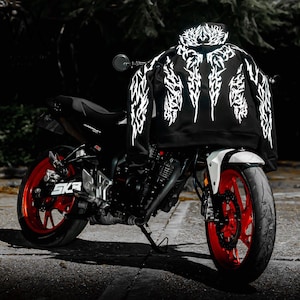 May include: A black hooded sweatshirt featuring a white, intricate, jagged tribal-style graphic pattern across the back and sleeves, draped over a black motorcycle with bright red wheels and a white front fender, set against a dark background.