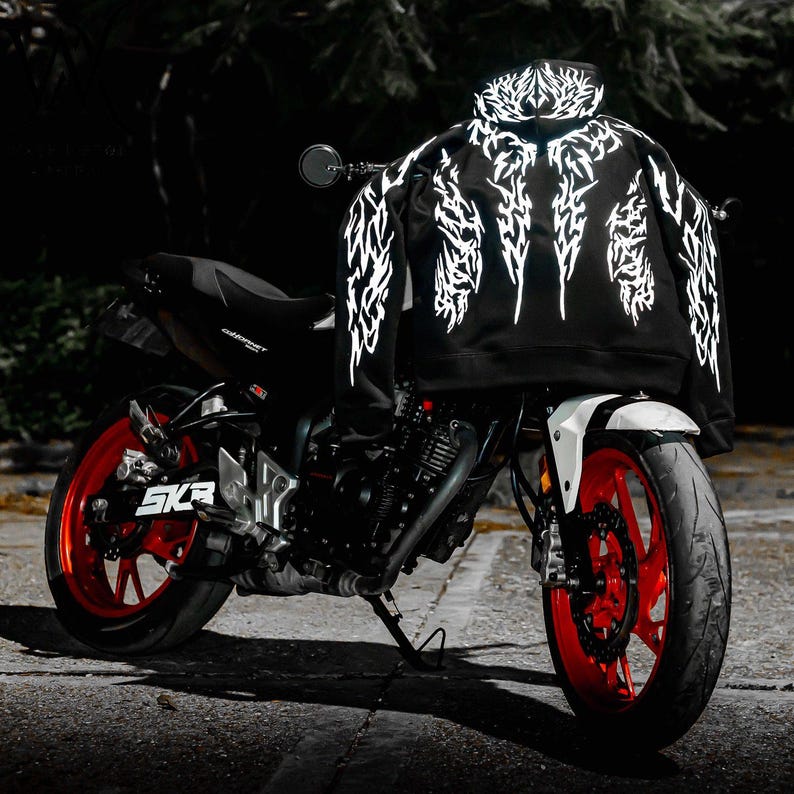 May include: A black hooded sweatshirt featuring a white, intricate, jagged tribal-style graphic pattern across the back and sleeves, draped over a black motorcycle with bright red wheels and a white front fender, set against a dark background.