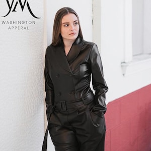 May include: A black double-breasted faux leather jumpsuit featuring a notched collar, long sleeves, and a matching belt with a rectangular buckle at the waist. The garment includes the text WASHINGTON APPERAL with a stylized logo in the upper corner.