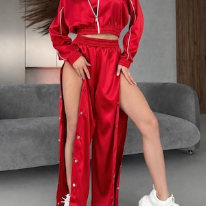 May include: A red satin tracksuit set. The set includes a cropped zip-up jacket with white trim, a matching crop top, and high-waisted trousers with snap closures along the sides. The outfit is paired with white trainers.