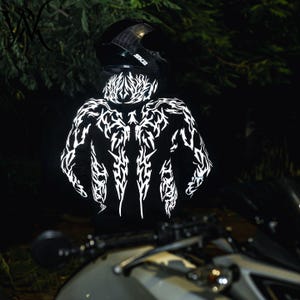 May include: A black hooded sweatshirt featuring a white reflective tribal-style graphic pattern across the back and hood. The garment is worn with a black motorcycle helmet, and the reflective design is illuminated against a dark background.