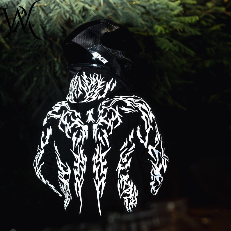 May include: A black hooded sweatshirt featuring a reflective white tribal-style graphic pattern across the back and hood, worn with a black motorcycle helmet. The design consists of sharp, jagged lines that contrast against the dark fabric.
