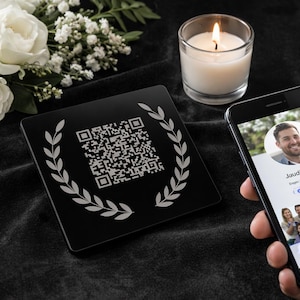 May include: A black memorial QR code plaque with a laurel wreath design, next to a lit candle and a smartphone displaying a profile. White roses and greenery are in the background.