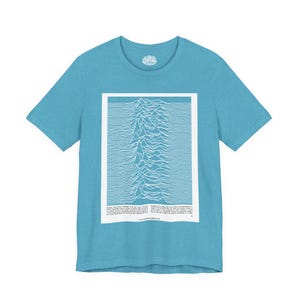 May include: A light blue t-shirt featuring a white graphic design. The graphic is a stylized mountain range with horizontal lines. The shirt has a crew neck and short sleeves. The text at the bottom of the graphic is difficult to read.