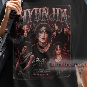 May include: A black short-sleeved t-shirt featuring a graphic print of the name HYUNJIN in a metallic font, multiple images of a person, the text Stray Kids, and five stars. The shirt is worn with black shorts and a black backpack.