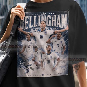May include: A black short-sleeved t-shirt featuring a graphic print of Jude Bellingham in an England football kit. The design includes the text BELLINGHAM, Jude Bellingham 10, and multiple images of the player in action poses on a blue background.