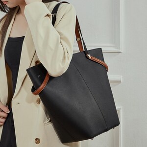 May include: Black tote bag with brown leather handles and accents. The bag has a structured design and a textured finish. The handles are attached with silver-toned hardware. The bag is carried over the shoulder.